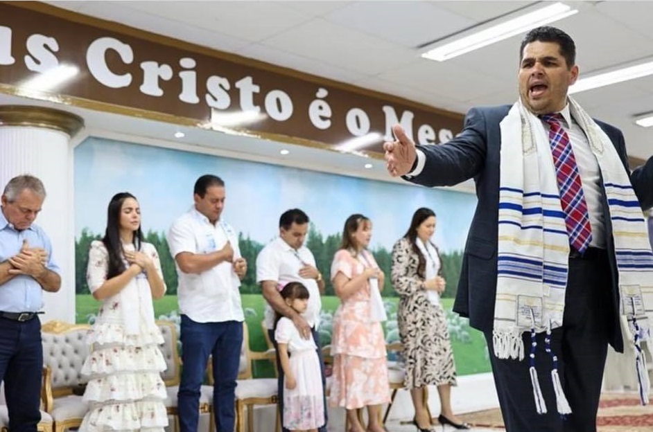 Weverton e Josimar e seus convidados com as mãos no peito sob a oração de um pastor da igreja...